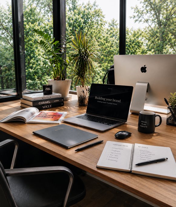 A sunlit desk by tall windows — a laptop reading "Building your brand," notebooks, and coffee